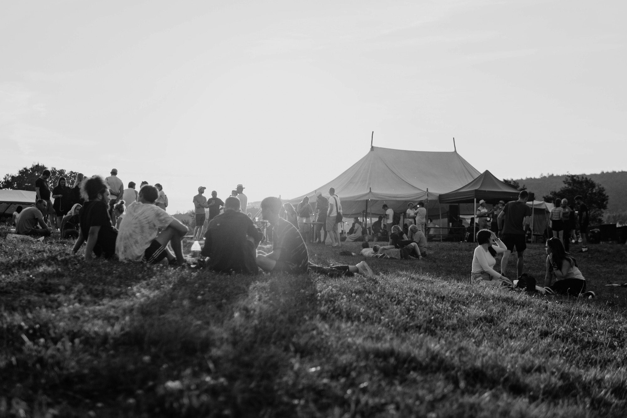 People sitting and standing on a grassy hillside beside a large marquee